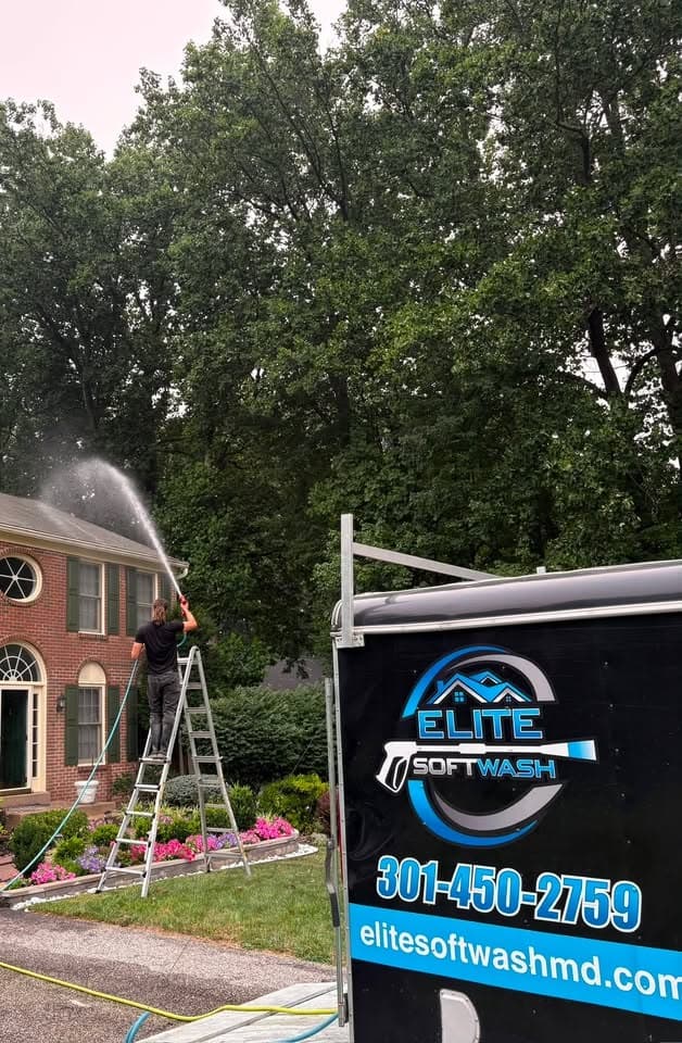 Elite Soft Wash technician on a ladder soft washing a brick colonial, branded trailer in the driveway