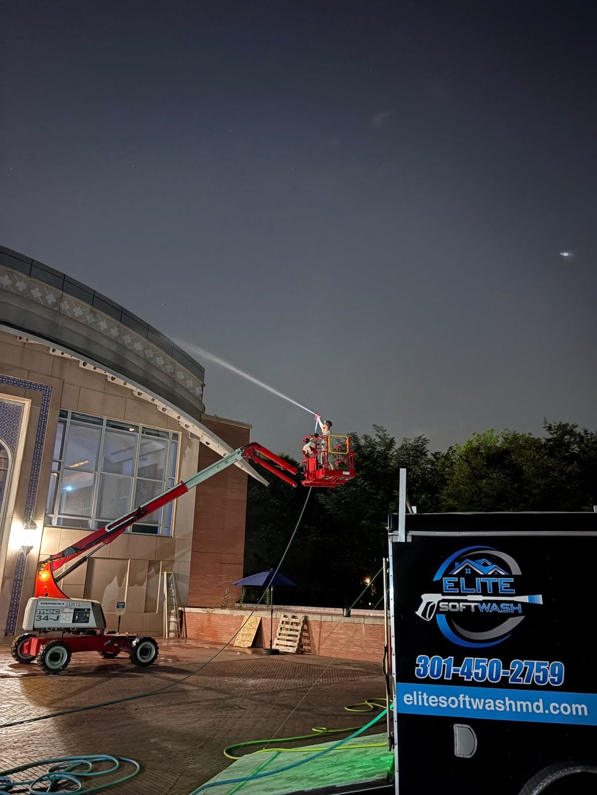Elite Soft Wash technician on an extended boom lift soft washing a curved stone facade at night, branded trailer on site